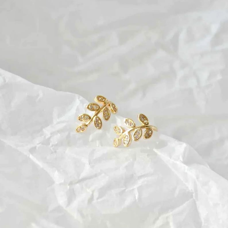 May include: A pair of gold-toned stud earrings shaped like delicate leaf branches. Each earring features a curved stem with multiple small, oval-shaped leaves, each embedded with tiny, clear, sparkling stones that catch the light.