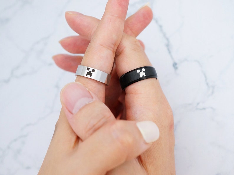May include: Two matching rings with a black and white pixelated creeper face design. The rings are worn on the index fingers of two hands.