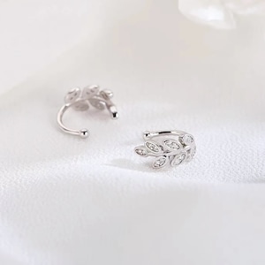May include: A pair of silver-toned ear cuffs featuring a delicate leaf-shaped design. Each cuff is adorned with small, clear, sparkling stones embedded along the surface of the leaves, creating a bright, reflective, and metallic appearance.