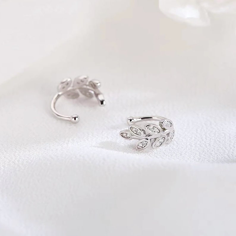 May include: A pair of silver-toned ear cuffs featuring a delicate leaf-shaped design. Each cuff is adorned with small, clear, sparkling stones embedded along the surface of the leaves, creating a bright, reflective, and metallic appearance.