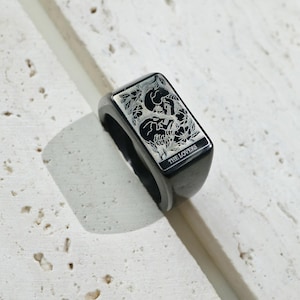 May include: A black rectangular signet ring featuring an engraved illustration of two skulls intertwined with floral elements. The engraving includes the text THE LOVERS at the bottom of the rectangular face, rendered in a clear, sans-serif font.