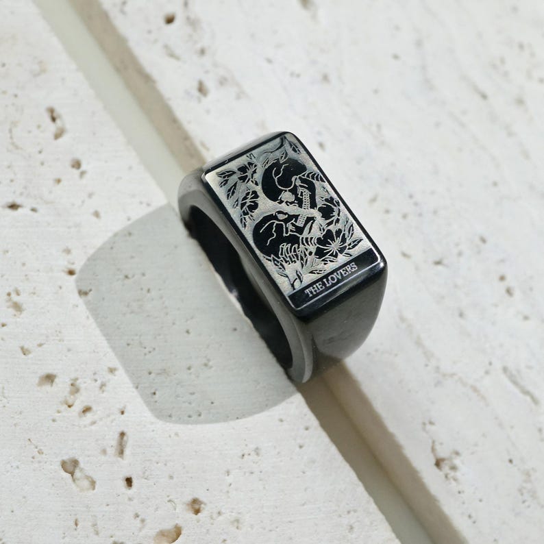 May include: A black rectangular signet ring featuring an engraved illustration of two skulls intertwined with floral elements. The engraving includes the text THE LOVERS at the bottom of the rectangular face, rendered in a clear, sans-serif font.