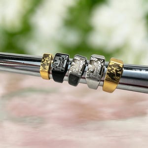 May include: Five rectangular signet rings arranged in a row, featuring engraved tarot card imagery. The collection includes gold, black, and silver-toned metal finishes, each displaying detailed illustrations of figures and celestial motifs.