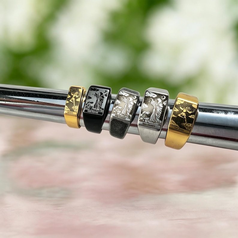 May include: Five rectangular signet rings arranged in a row, featuring engraved tarot card imagery. The collection includes gold, black, and silver-toned metal finishes, each displaying detailed illustrations of figures and celestial motifs.