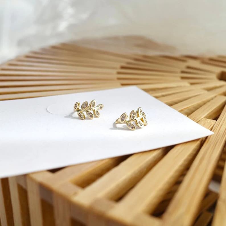 May include: A pair of gold-toned ear cuffs shaped like small leafy branches. Each piece features a series of marquise-shaped leaves, with each leaf inlaid with a small, clear, sparkling stone. The jewelry is displayed on a white card.