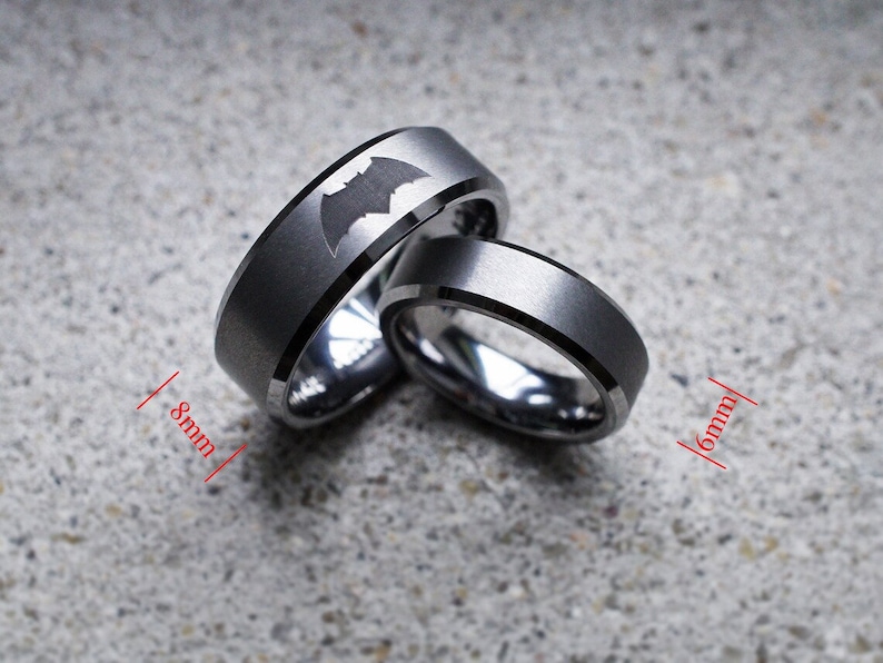 May include: Two tungsten carbide rings with a black matte finish. The wider ring is 8mm wide and features a Batman logo. The narrower ring is 6mm wide.