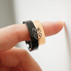 Könnte beinhalten: Zwei ineinandergreifende Ringe, einer schwarz und einer goldfarben, mit einem Design aus zwei Händen, die ein Herz mit den Buchstaben C und M darin halten. Die Ringe werden an einem Finger getragen.
