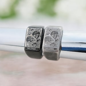 May include: Two rectangular signet rings featuring an engraved tarot card design titled THE LOVERS. One ring is finished in a polished black tone, while the other is in a brushed silver tone. Both rings display a detailed illustration of two skulls.