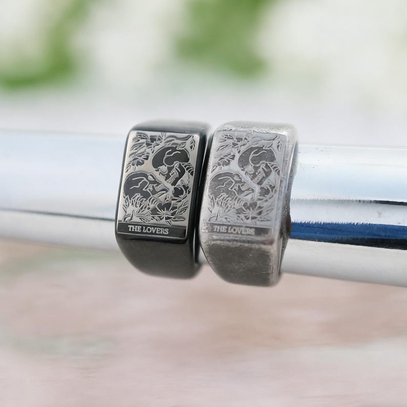 May include: Two rectangular signet rings featuring an engraved tarot card design titled THE LOVERS. One ring is finished in a polished black tone, while the other is in a brushed silver tone. Both rings display a detailed illustration of two skulls.