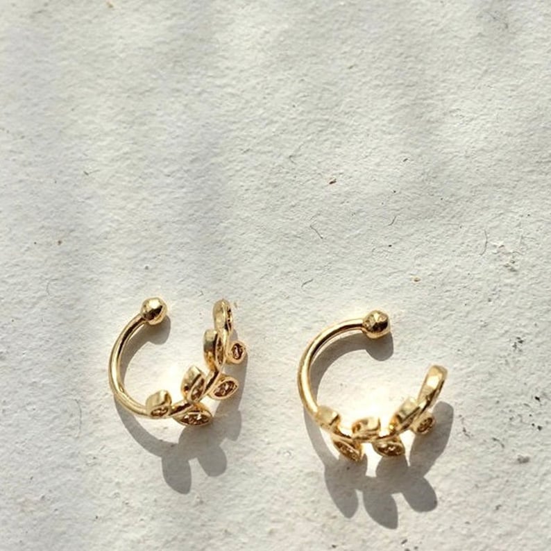 May include: A pair of gold-toned ear cuffs featuring a delicate leaf-inspired design. Each piece is shaped as a curved band with small, interconnected leaf motifs along one side and a smooth, rounded terminal at the top of the open hoop structure.