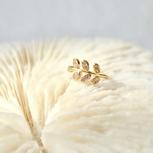 May include: A gold-toned ear cuff shaped like a delicate branch with five small leaves. Each leaf is embellished with a tiny, clear, sparkling stone. The piece features a slender, curved metal band designed to wrap around the edge of the ear.
