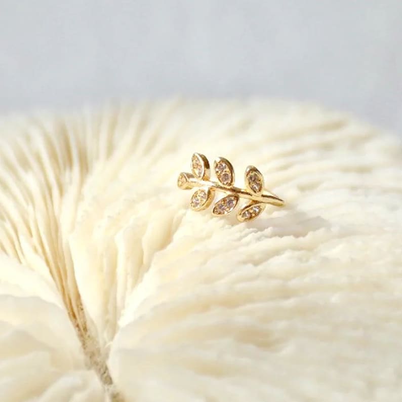 May include: A gold-toned ear cuff shaped like a delicate branch with five small leaves. Each leaf is embellished with a tiny, clear, sparkling stone. The piece features a slender, curved metal band designed to wrap around the edge of the ear.