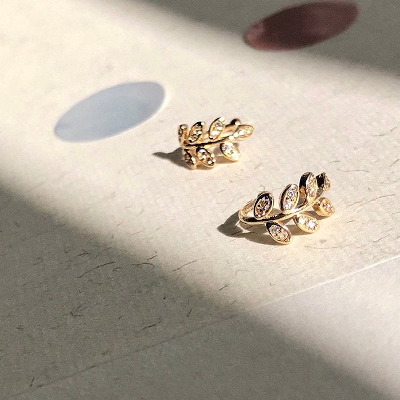 May include: A pair of gold-toned ear cuffs shaped like delicate leafy branches. Each piece features a curved metal stem adorned with small, clear, sparkling stones set into individual leaf-shaped segments, creating a botanical design.