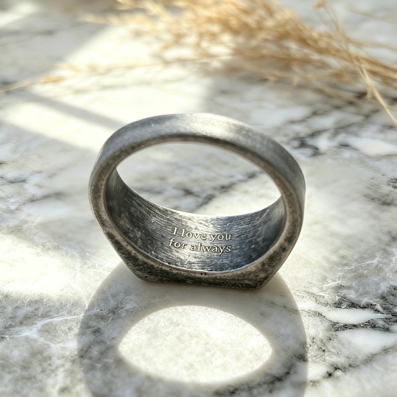 May include: A dark, weathered metal ring with a textured, matte finish. The interior of the band features a clear, engraved inscription that reads, "I love you for always," centered within the circular opening of the band.