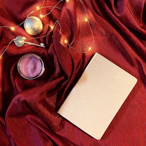 Puede incluir: Un cuaderno blanco con una cubierta en blanco se encuentra sobre un fondo de tela roja con una vela, una llave y una cadena de luces.