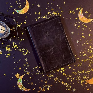 Puede incluir: Un diario encuadernado en cuero negro con detalles dorados sobre un fondo azul oscuro rodeado de purpurina dorada y estrellas.