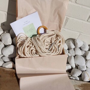 May include: A kit for making a macrame plant hanger. The kit includes natural cotton rope, a wooden ring, and a metal hook. The kit is packaged in a light pink box with tissue paper.