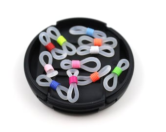 May include: A collection of clear plastic eyeglass temple tip covers with colorful rubber bands. The covers are in a black round container.