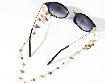 May include: Black sunglasses with a gold chain featuring purple gemstone beads. The chain is attached to the arms of the sunglasses.