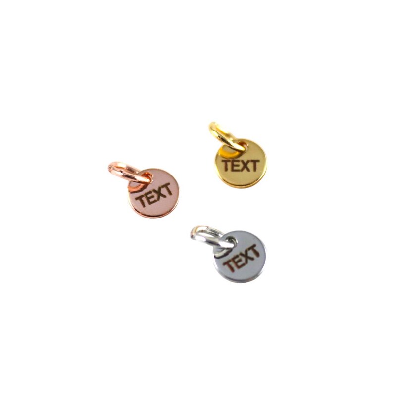 May include: Three small round metal charms with a loop at the top. Each charm is engraved with the word "TEXT". The charms are gold, rose gold, and silver.