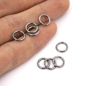 May include: A close-up of six black metal jump rings. The rings are different sizes and are arranged in a random pattern on a white surface.