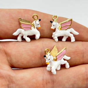 May include: Three white unicorn charms with gold wings and pink accents. Each charm has a gold loop at the top for attaching to jewelry. The unicorns have black horns and orange noses.