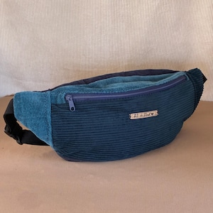 Peut inclure: Un sac banane en velours côtelé avec un corps principal bleu foncé et une poche avant bleu sarcelle. Le sac comprend une fermeture éclair bleu foncé, une sangle noire réglable avec une boucle en plastique et une étiquette "lili chillout".