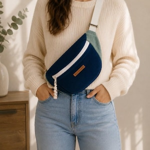 Peut inclure: Sac banane en velours côtelé bleu marine avec fermeture éclair et sangle blanches. Le sac comporte une petite étiquette en cuir marron et un pompon blanc. Accessoire de mode porté en bandoulière.