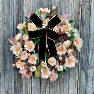 May include: A circular floral wreath featuring peach-colored flowers, buds, and leaves, accented by a dark brown velvet bow with gold trim. The wreath is set against a weathered wooden door.