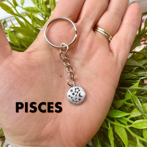 May include: Silver keychain with a round charm displaying the Pisces constellation, stars, and a crescent moon. The word "PISCES" is printed in black. The keychain is attached to a silver ring.