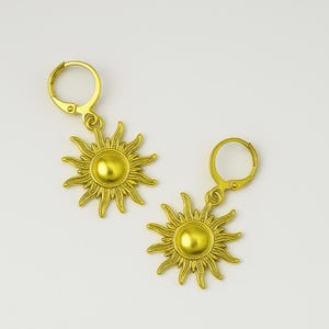 Op de afbeelding: Een paar goudkleurige oorbellen in de vorm van een zon. Elke oorbel heeft een gedetailleerd zonnesterontwerp met een centraal cirkelvormig element en een beugelsluiting. De oorbellen worden tegen een witte achtergrond getoond.