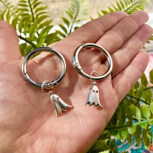 May include: Two silver keychains with ghost charms. Each keychain has a large, round, silver ring with a small ghost charm hanging from it. The ghost charms are silver and have two small eye holes and a wavy bottom edge.