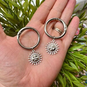 May include: Two silver-colored hoop earrings with a sun charm. Each charm features a smiling sun face with radiating rays. The earrings are held in a hand against a background of green foliage.