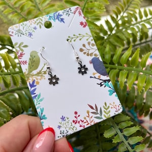 May include: A pair of silver dangle earrings with a black flower charm. The earrings are displayed on a white card with a floral design.