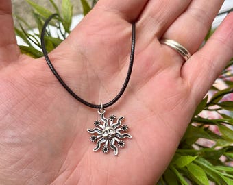 May include: A silver-colored sun pendant necklace with a black cord. The sun pendant has a smiling face in the center, with rays and small black accents. The necklace is displayed on a hand.