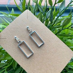 Puede incluir: Un par de pendientes rectangulares plateados. Cada pendiente presenta una pequeña bola plateada en la parte superior, conectada a un marco rectangular. Los pendientes se exhiben sobre una tarjeta marrón, con follaje verde de fondo.