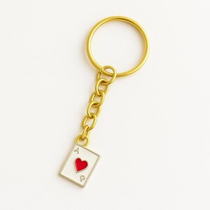 May include: Gold-coloured keychain with a playing card charm. The charm is a white card with a red heart and the letter "A". The keychain has a ring and chain design.