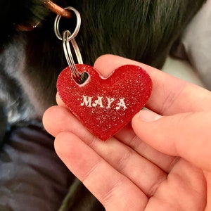 May include: Red heart-shaped dog tag with the name "MAYA" in white lettering. The tag is attached to a silver key ring.