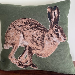 Puede incluir: Una almohada verde con una imagen bordada de una liebre corriendo, en tonos marrones y blancos.