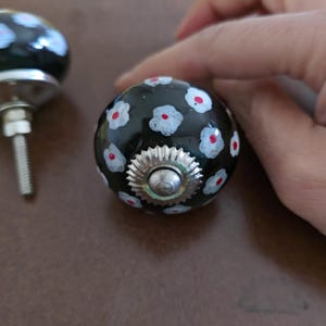 Puede incluir: Dos pomos de armario de cer&aacute;mica negra con dise&ntilde;os florales blancos y rojos. Los pomos tienen una base de metal plateado.