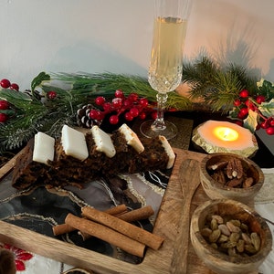 May include: A wooden serving tray with a black and white agate design. The tray features a slice of fruitcake topped with white icing, cinnamon sticks, a wooden spoon, and two small wooden bowls filled with pecans and raisins. The tray is set on a white tablecloth with a sprig of holly and a glass of champagne.