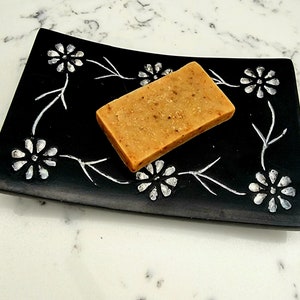 Könnte beinhalten: Eine Seife auf einem schwarzen Seifenschälchen mit floralem Muster. Die Seife ist hellbraun und hat eine raue Textur.