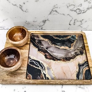 May include: A wooden serving tray with two small wooden bowls. The tray has a black, brown, and gold abstract design.