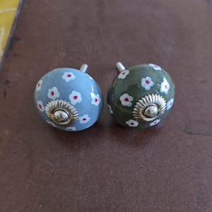 Puede incluir: Dos pomos de caj&oacute;n de cer&aacute;mica con dise&ntilde;o floral. Un pomo es azul claro con flores blancas y centros rojos. El otro pomo es verde con flores blancas y centros rojos.
