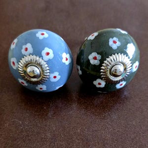 Puede incluir: Dos pomos de caj&oacute;n de cer&aacute;mica con un dise&ntilde;o floral. Un pomo es azul con flores blancas y centros rojos. El otro pomo es verde con flores blancas y centros rojos.