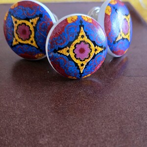 Puede incluir: Tres pomos redondos de cer&aacute;mica para armario con un patr&oacute;n geom&eacute;trico azul, rojo, amarillo y rosa. Los pomos tienen una base blanca.