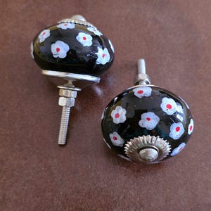 Puede incluir: Dos pomos de armario de cer&aacute;mica negra con dise&ntilde;os florales blancos y rojos. Los pomos tienen una base de metal plateado y un tornillo.