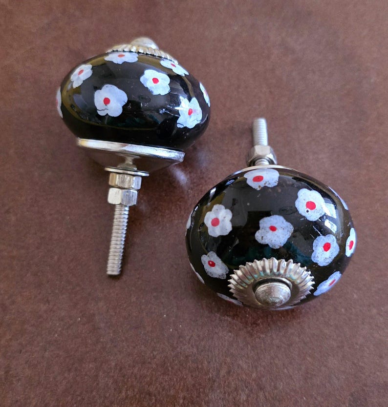 Puede incluir: Dos pomos de armario de cer&aacute;mica negra con dise&ntilde;os florales blancos y rojos. Los pomos tienen una base de metal plateado y un tornillo.