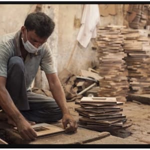 May include: A person wearing a face mask is working on a wooden frame in a workshop. The frame is made of several pieces of wood that have been cut and joined together. There are stacks of similar wooden frames in the background.