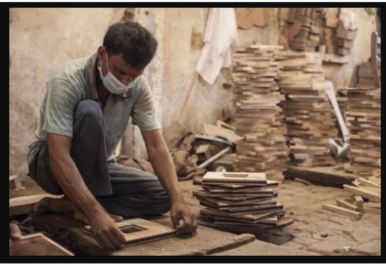 May include: A person wearing a face mask is working on a wooden frame in a workshop. The frame is made of several pieces of wood that have been cut and joined together. There are stacks of similar wooden frames in the background.